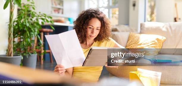 Kvinna jämför dokument med information på en surfplatta i ett ljust vardagsrum.